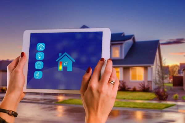 Eine Person steht vor einem Haus und hält ein Touchpad, auf dem u.a. Temperatur, Licht und Musik eingestellt werden können. 