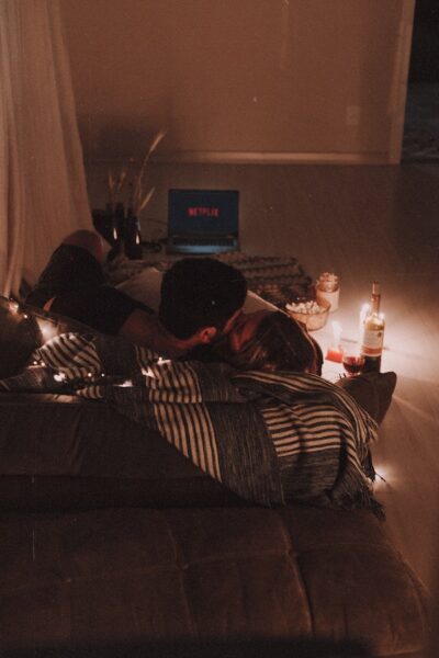 Ein Paar küsst sich auf einem Sofa vor einem Laptop, auf dem Netflix spielt. Neben ihnen stehen Kerzen, Wein und Popcorn. 