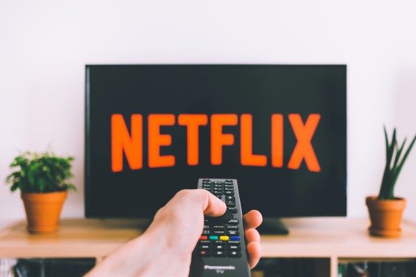 Eine Person hat eine Fernbedienung in der linken Hand und richtet diese auf einen Bildschirm, auf dem das Netflix-Logo zu sehen ist. 