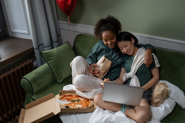 Zwei Frauen kuscheln auf einem Sofa mit Laptop, Pizza, Popcorn und Decken