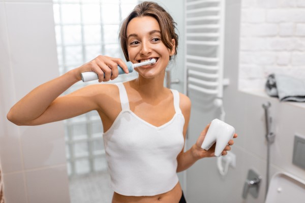 Eine Frau steht im Bad und putzt sich die Zähne mit einer elektrischen Zahnbürste. In der anderen Hand hält sie eine Zahnmodell.