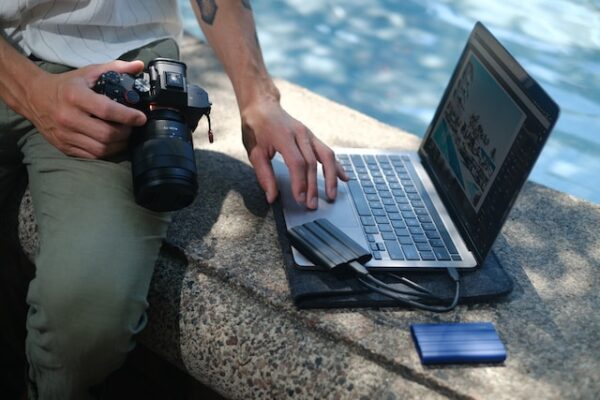 Eine Person sitzt im Freien. Sie nutzt einen Laptop, während sie eine Kamera auf dem Schoß hält.