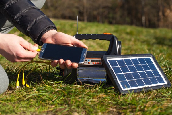Eine Person ist im Freien und steckt ein Kabel an ein Smartphone. Das Kabel kommt aus einer Powerbank, die durch ein kleines Solarpanel beladen wird. 